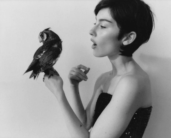 Profile shot of a short-haired woman holding a small owl perched on her hand against a plain white background.