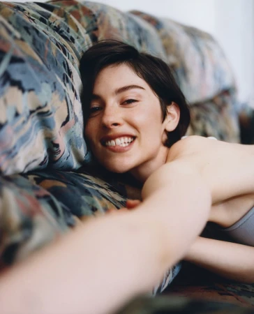 Close-up portrait of a smiling woman with short dark hair reclining against a patterned floral couch, photographed by Laura Jane Coulson.