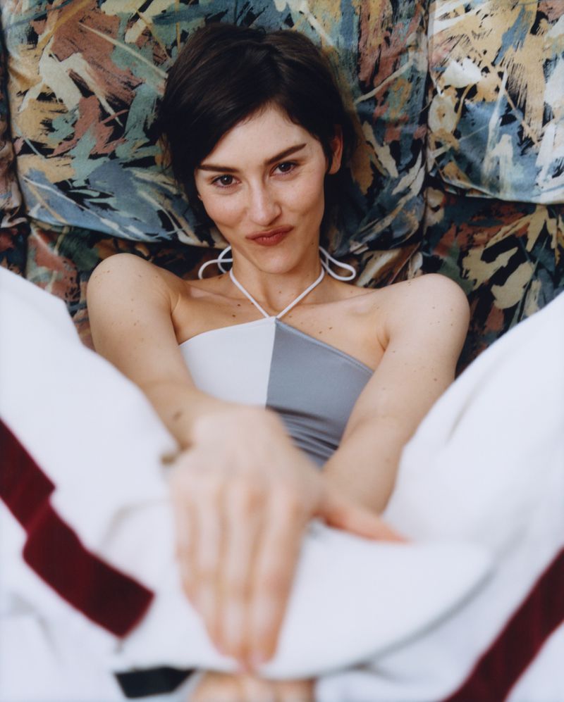 Portrait of a woman with short dark hair wearing a white and grey halter top, seated against an abstract painted backdrop