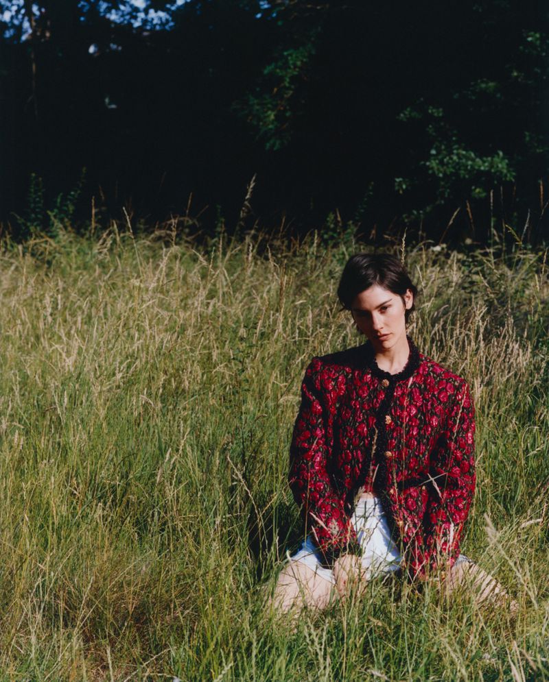 Woman wearing a patterned red jacket kneeling in a tall grassy field outdoors under natural daylight.