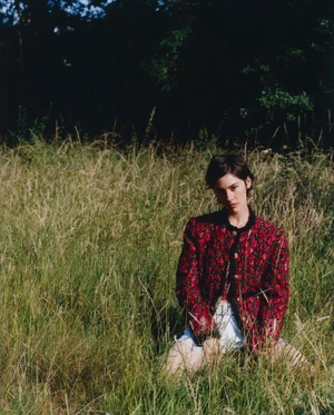 Woman wearing a patterned red jacket kneeling in a tall grassy field outdoors under natural daylight.