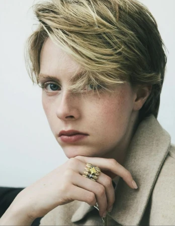 Close-up portrait of a woman with short blonde hair wearing a beige jacket and a jewelled ring, against a plain white background.