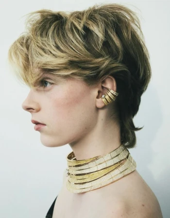 Profile portrait of a model wearing elaborate gold jewelry including a multi-strand choker and ear cuff against a white background