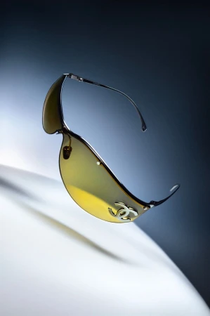 Close-up product shot of futuristic yellow-tinted sunglasses with a silver logo against a gradient background.