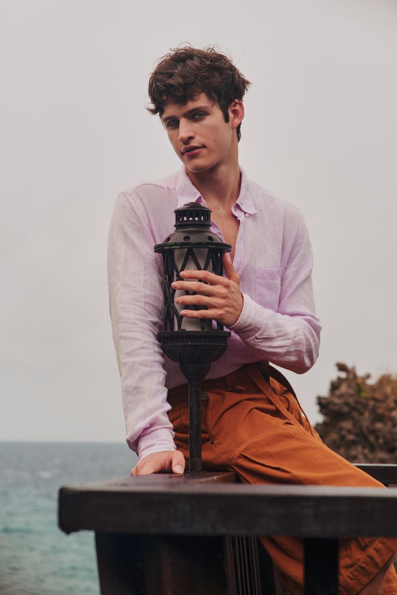 Young man in a pink shirt and rust trousers holding a black lantern on a balcony overlooking the ocean for Flanelle Magazine.