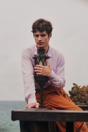A young man in a light purple shirt and brown trousers holding a decorative lantern against an overcast sky and ocean backdrop.