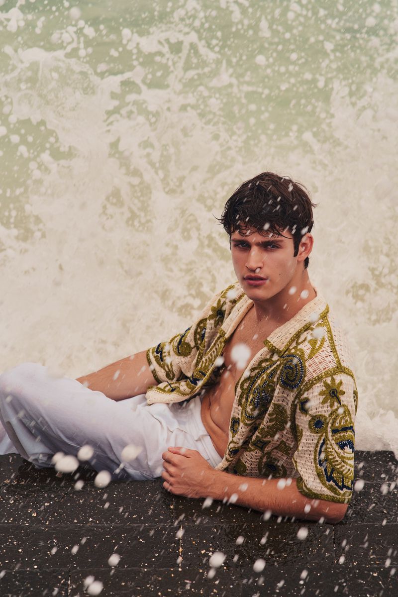 A man in an embroidered patterned shirt and white trousers lounging near crashing ocean waves, captured in a portrait by Anton Patdu for Flanelle Magazine.