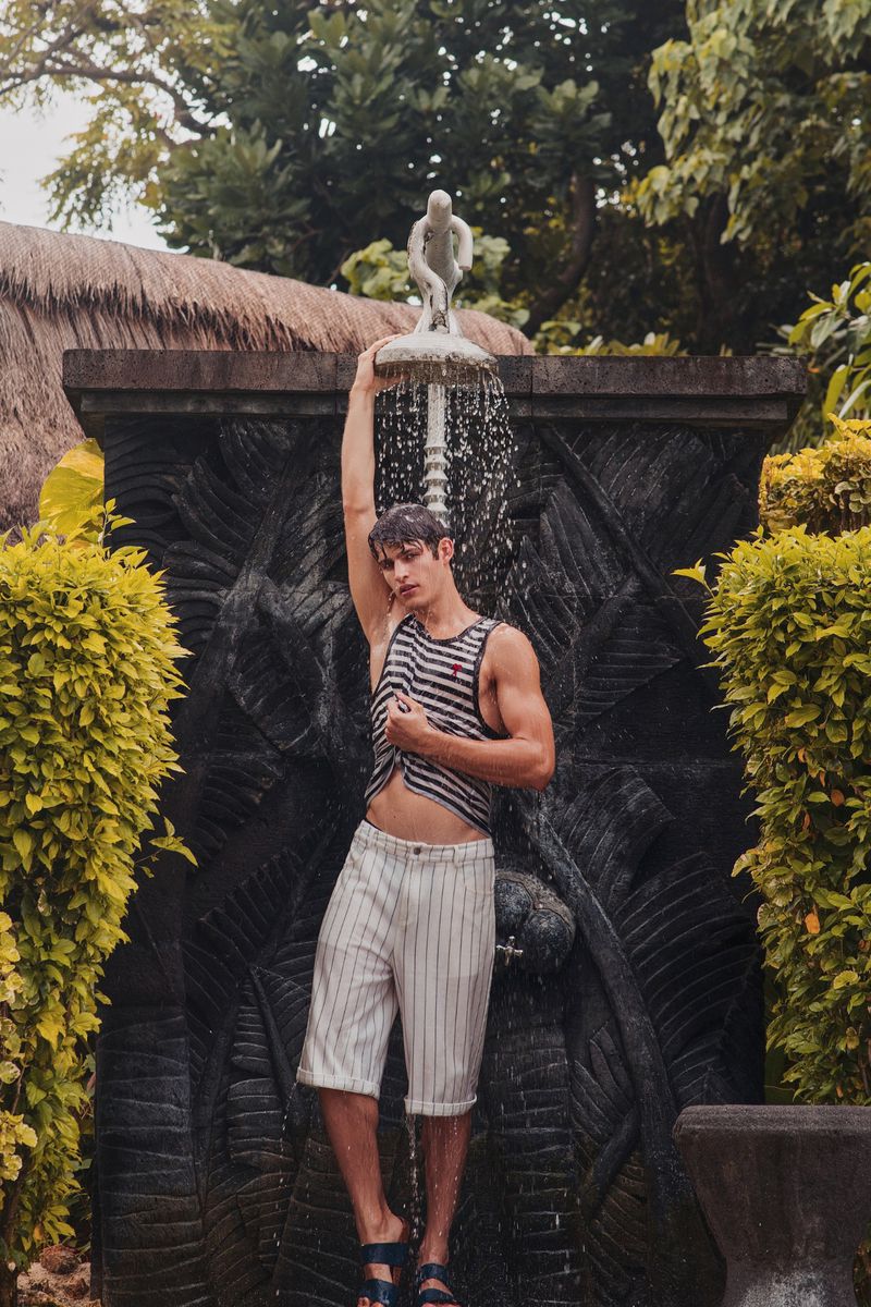 Man wearing a striped tank top and white trousers standing under an outdoor shower surrounded by lush foliage.