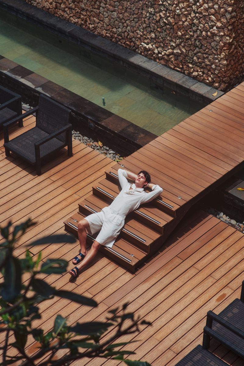 Man lying on wooden outdoor deck stairs next to a narrow pool wearing white shorts and a white top
