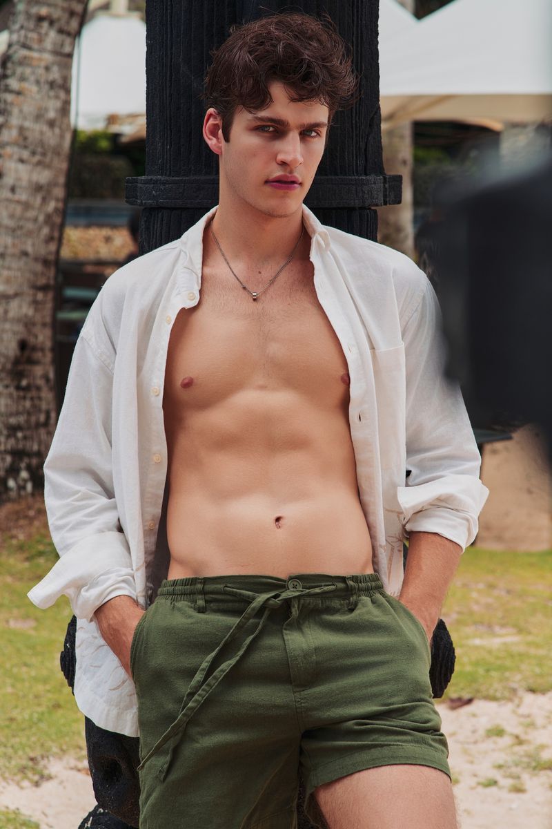 A young male model wearing an unbuttoned white shirt and olive green shorts leaning against a dark post outdoors.