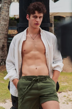 A young male model wearing an unbuttoned white shirt and olive green shorts leaning against a dark post outdoors.