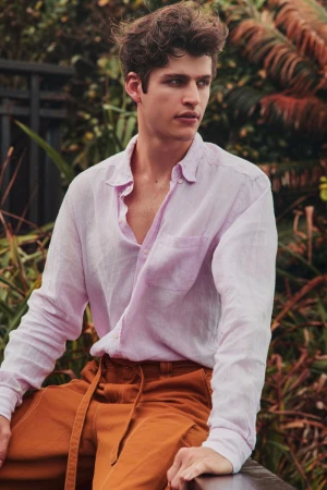 Young man with curly brown hair wearing a light pink linen shirt and rust-colored trousers outdoors amidst lush green foliage.