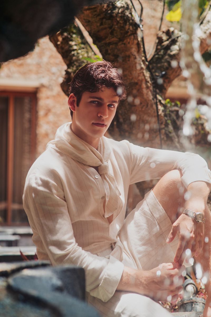 Young man in a white linen shirt and scarf seated outdoors near flowing water for Flanelle Magazine