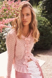 Model wearing a pink knit cardigan with bow details posing outdoors near pink flowers and green foliage