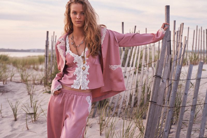 Model in pink silk pajamas with lace trim standing outdoors near a weathered wooden fence on a sandy beach at sunset.