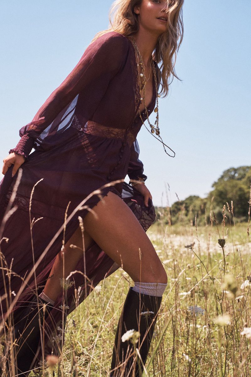 Model in long plum sheer dress wearing dark tall boots walking through a sunlit grassy meadow in a fashion campaign