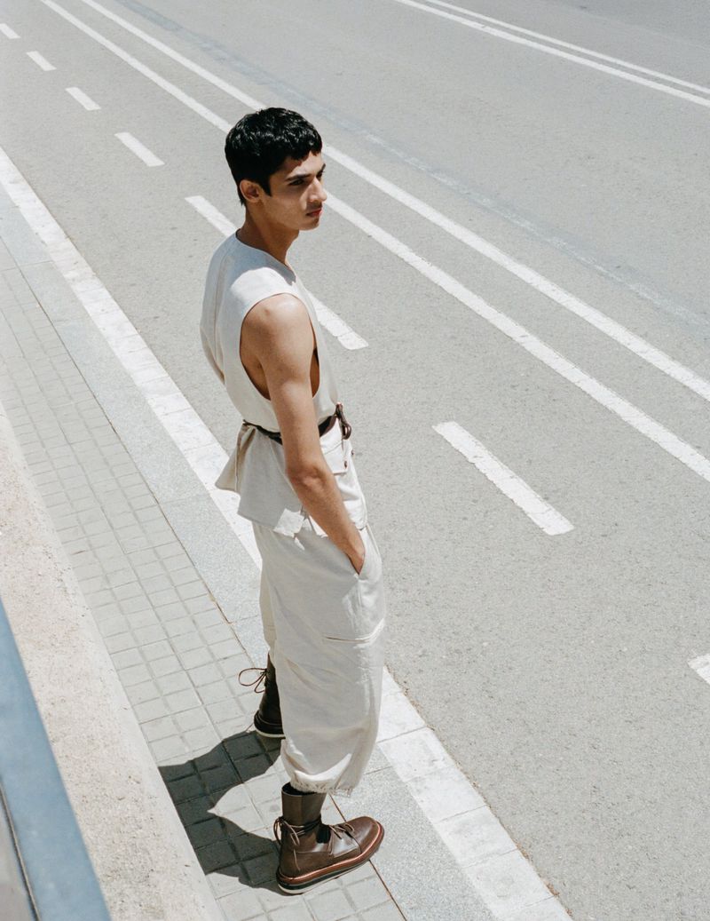Young adult man in sleeveless top and baggy pants standing on sidewalk beside road with painted white stripes in soft midday light.