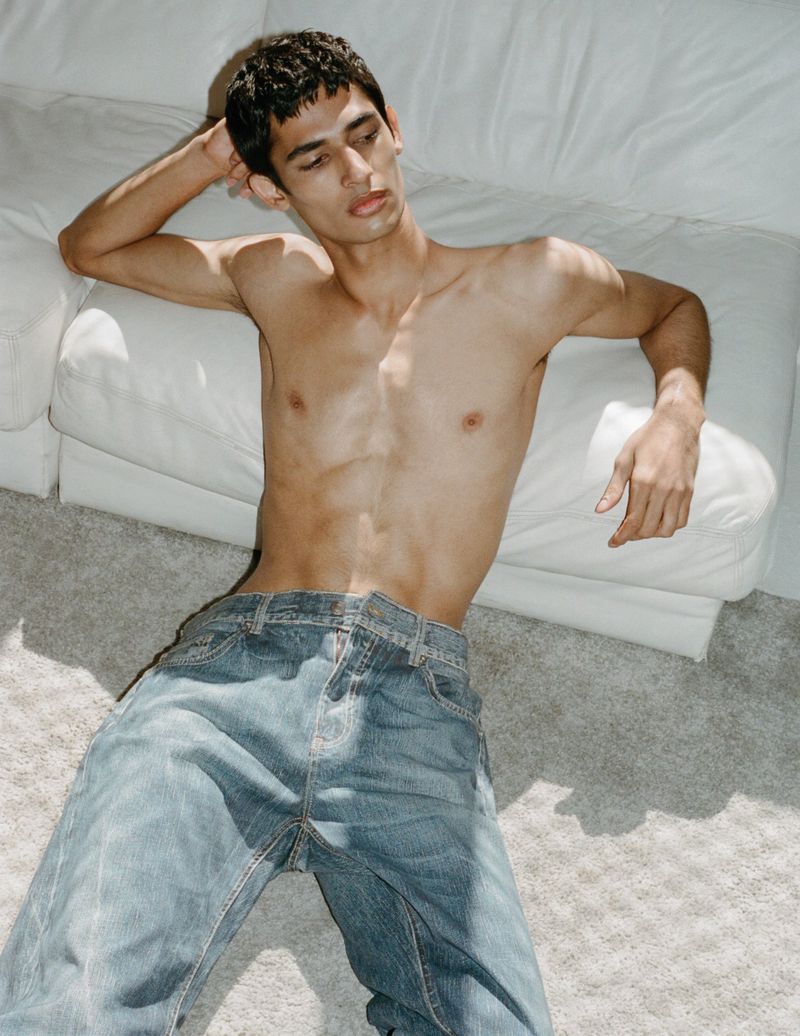 Topless man in light blue denim pants leans back on white sofa on gray carpet in indoor setting with bright, natural light.