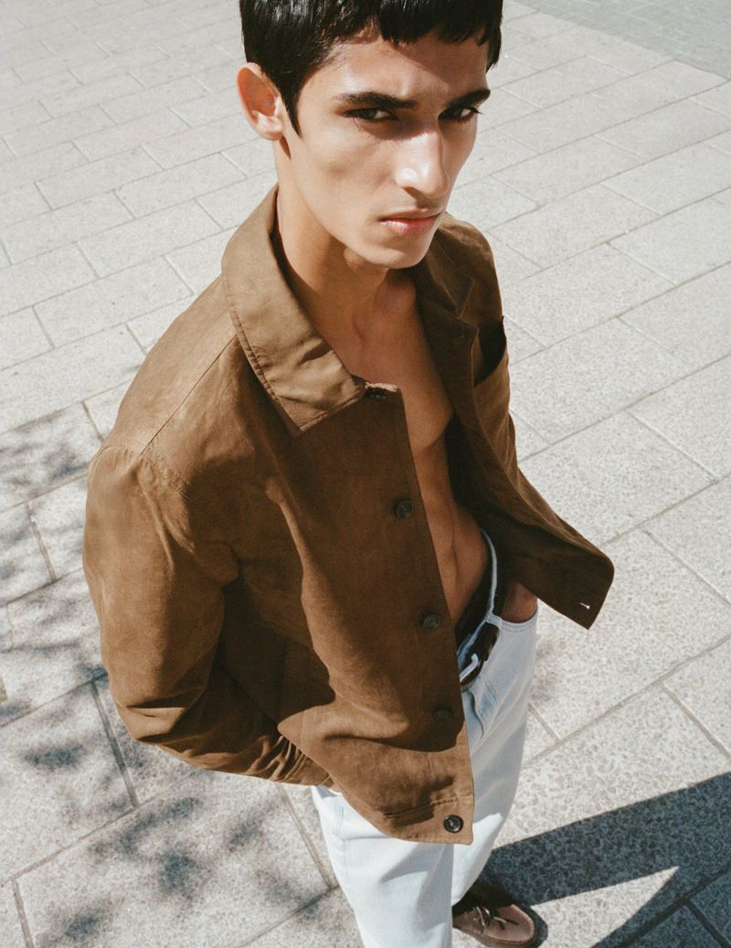 Model Nakul Bhardwaj wearing an open brown suede jacket and light trousers, looking up towards the camera on stone pavement.