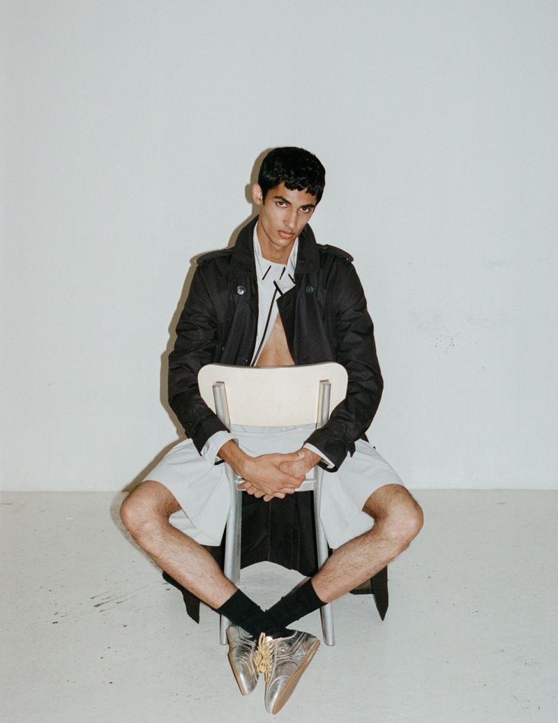 Young adult male fashion model posing on a chair in an indoor photo studio wearing a black trench coat, white shirt, and silver loafers.