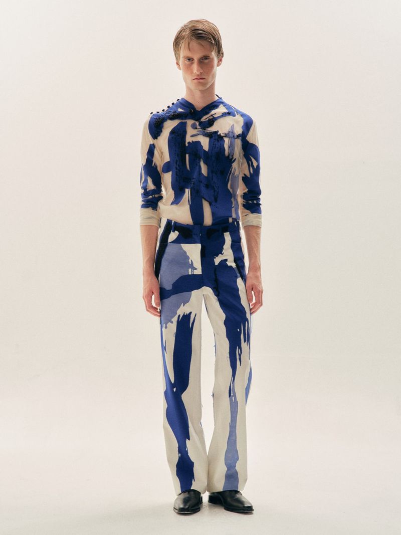 Full-length portrait of a male model wearing blue and white painterly print garments against a plain white studio backdrop.