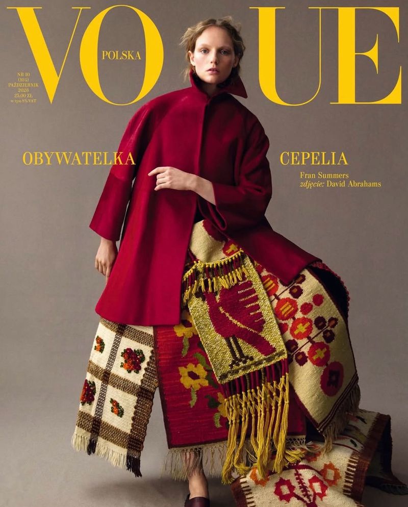 Model Fran Summers wearing a red coat and folk-patterned skirt on a grey seamless background for Vogue Polska.