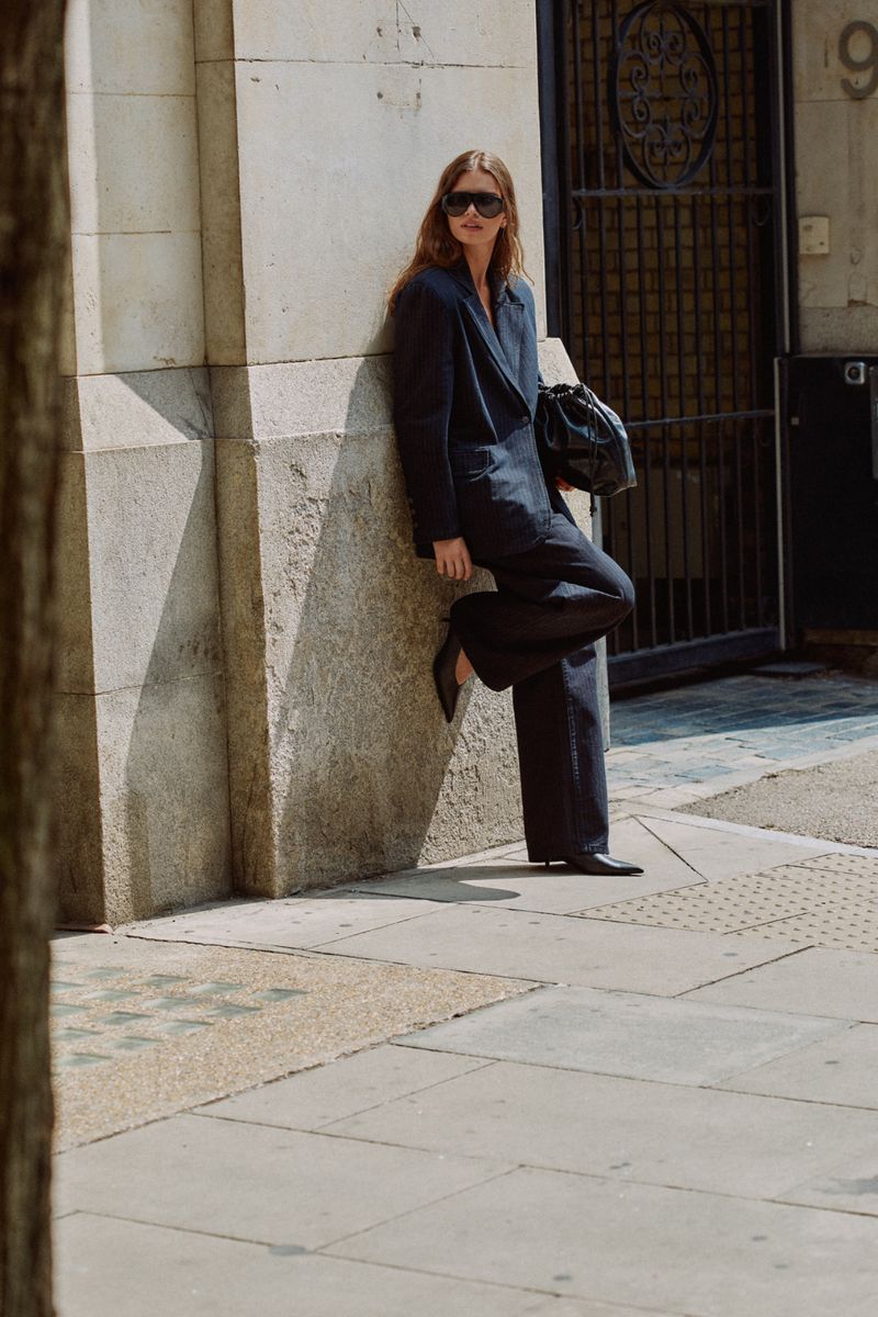 Woman in dark suit leaning against a stone building holding a black handbag outdoors