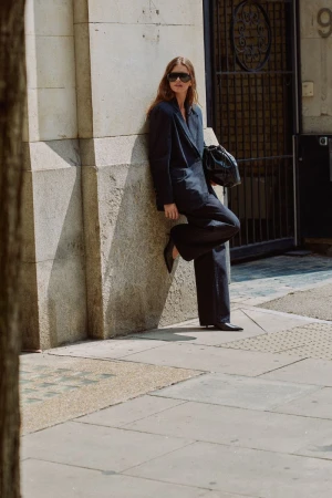 Woman in dark suit leaning against a stone building holding a black handbag outdoors