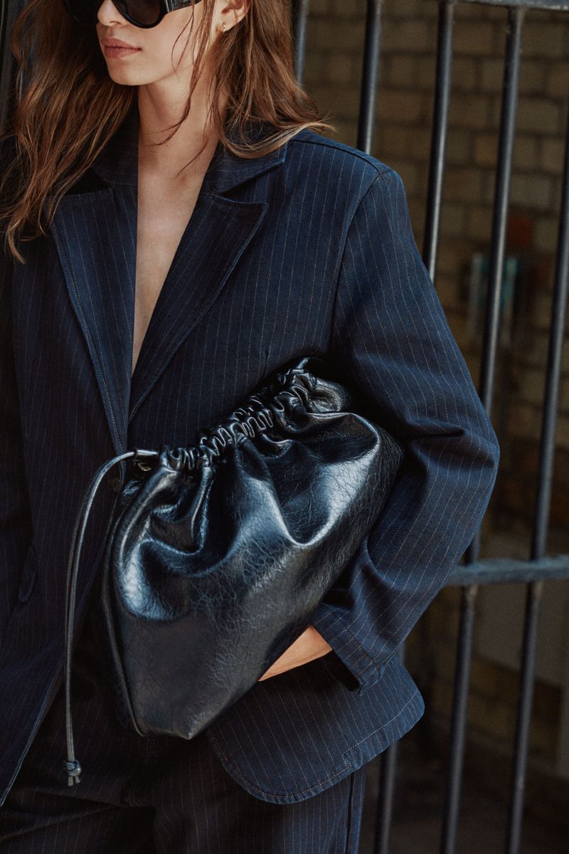 Model wearing dark sunglasses and a navy pinstripe blazer holding a black leather clutch bag against a black metal fence