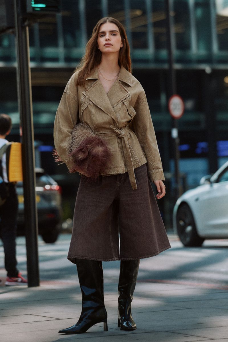 Woman wearing a beige jacket, brown culottes, and knee-high boots holding a textured pouch on a city street.