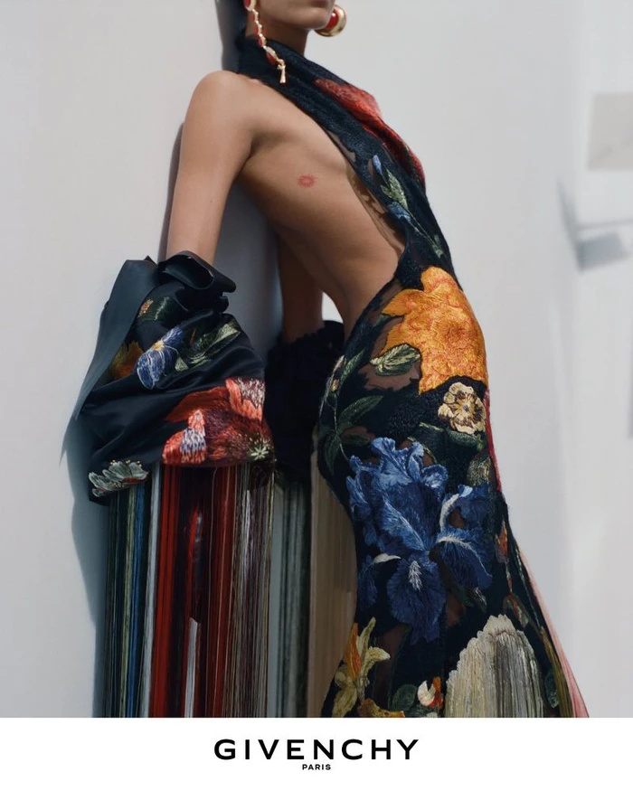 Model wearing a black gown with vibrant floral embroidery leaning against a plain white wall for Givenchy Winter 2026 campaign.