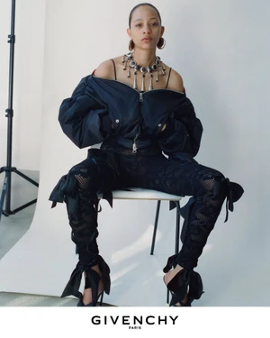 Model seated on a chair wearing a dark off-the-shoulder bomber jacket and patterned trousers against a white seamless backdrop