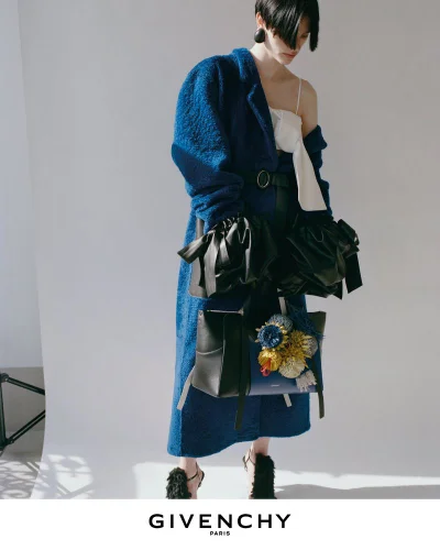 Model wearing a blue textured coat and carrying black handbags against a plain white studio backdrop.