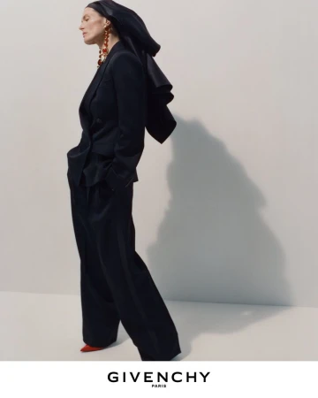 Model in black suit and hooded headwear posing against a plain backdrop with visible shadow for Givenchy Winter 2026 campaign.