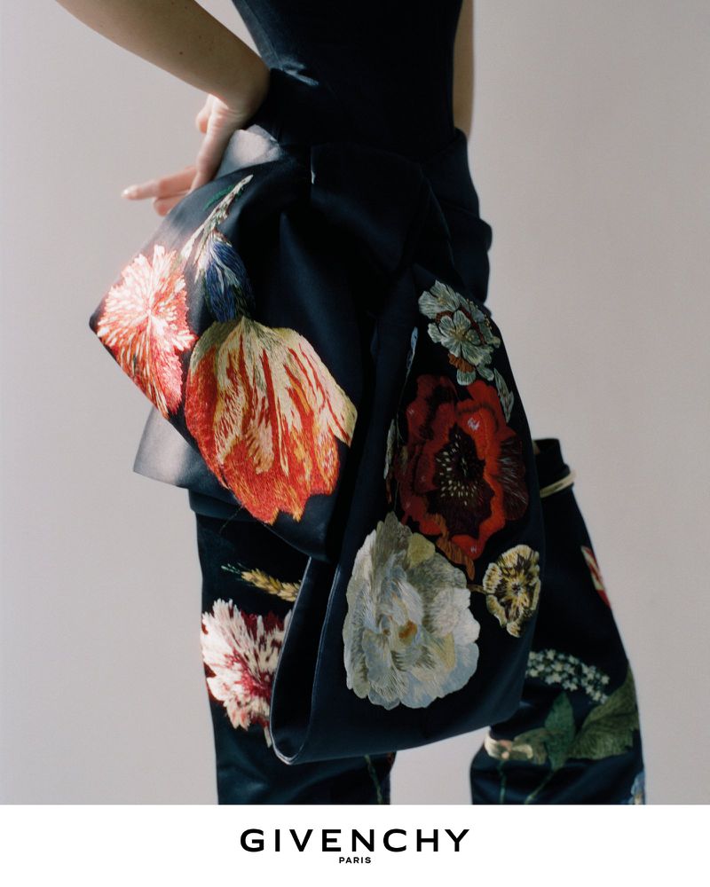 Close-up of a dark outfit featuring vibrant floral embroidery and a prominent bow detail against a neutral background.