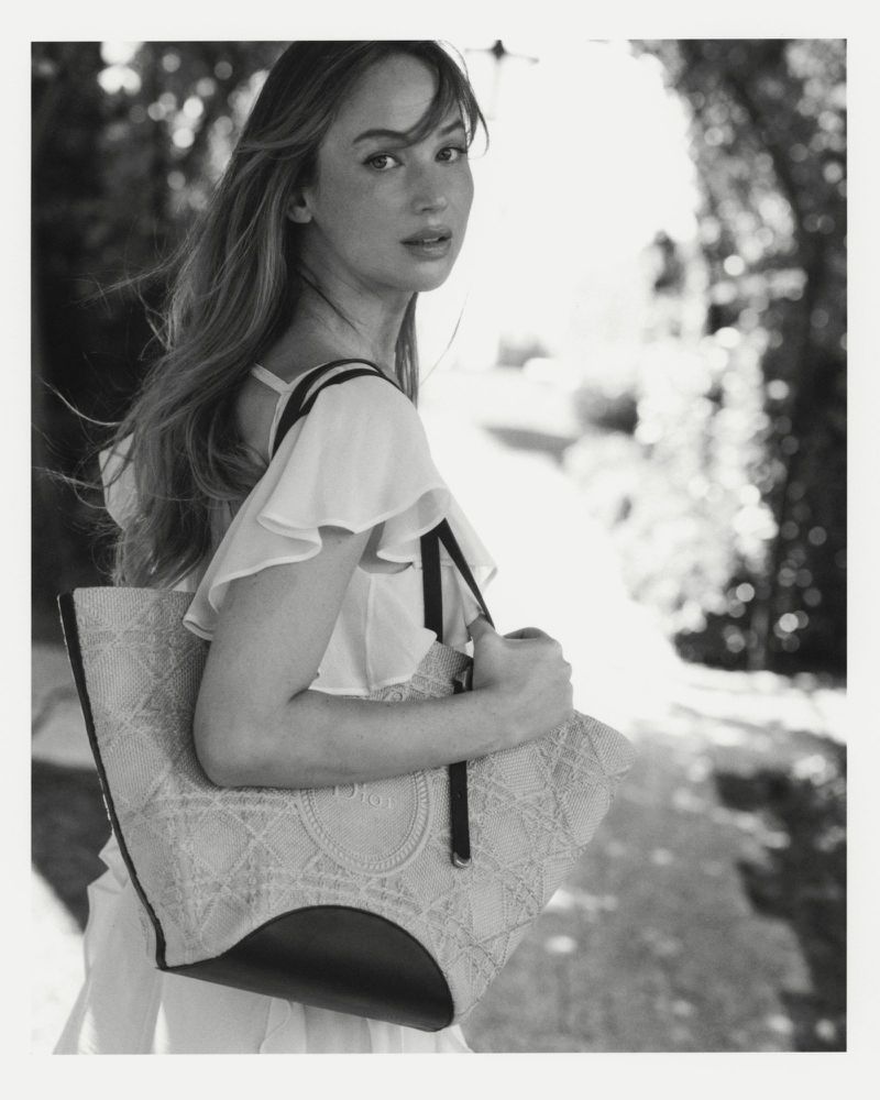 Jennifer Lawrence carrying a patterned Dior tote bag outdoors in a black and white portrait.