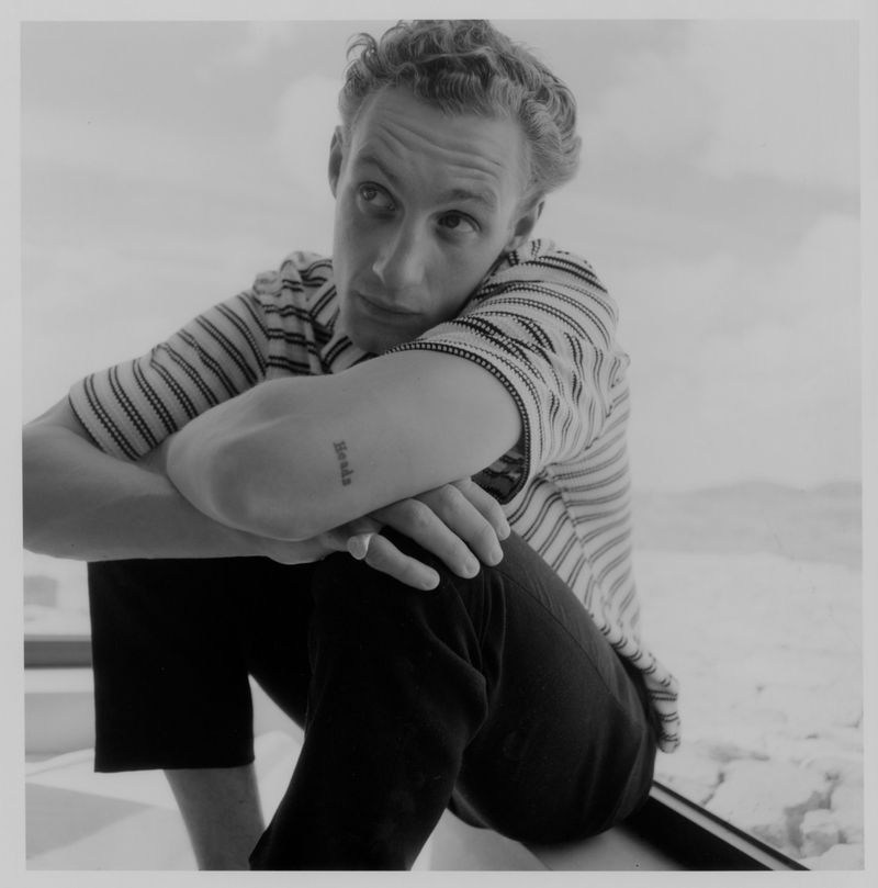 Black and white portrait of a young adult man in a striped shirt, sitting and looking away with a tattoo on his arm.