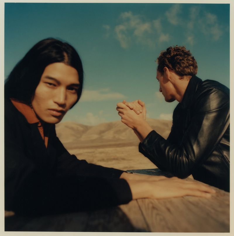 Two men seated at a wooden outdoor table in an arid desert landscape, one looking forward and the other in a black leather jacket.