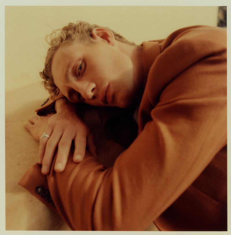 Close-up portrait of a young adult man in a brown suit resting his head on his arms, set against a neutral-toned background.