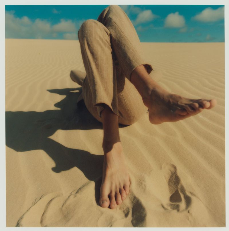 Person reclines on tan sand dunes with sunny blue sky. Lower legs and feet closest to camera, tan linen trousers on upper body.