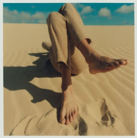 Person reclines on tan sand dunes with sunny blue sky. Lower legs and feet closest to camera, tan linen trousers on upper body.