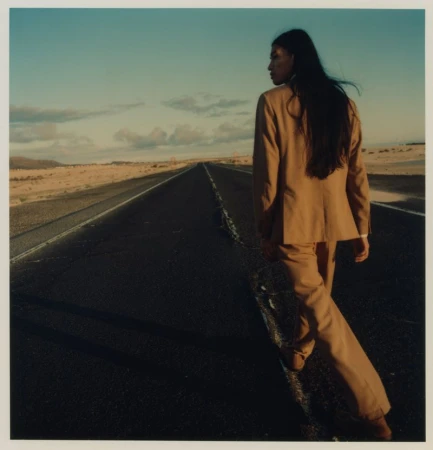 Woman in a tan suit walks down a desolate asphalt road under a blue sky. The desert stretches into the horizon under daylight.