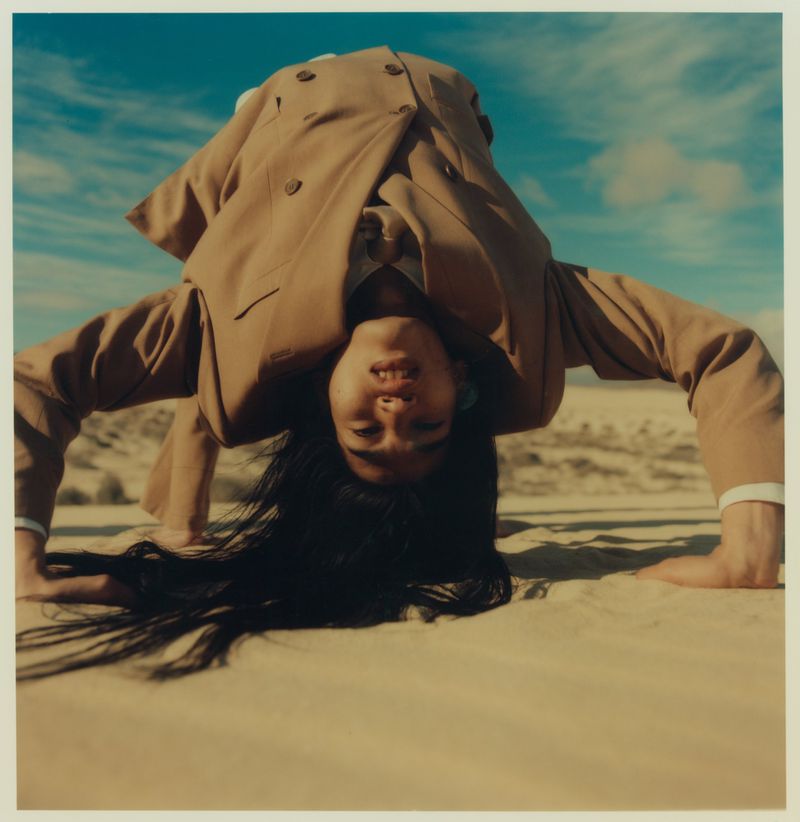 A person in a tan suit is doing a handstand on sand in a desert landscape under a blue sky with clouds, creating an unusual and striking visual.