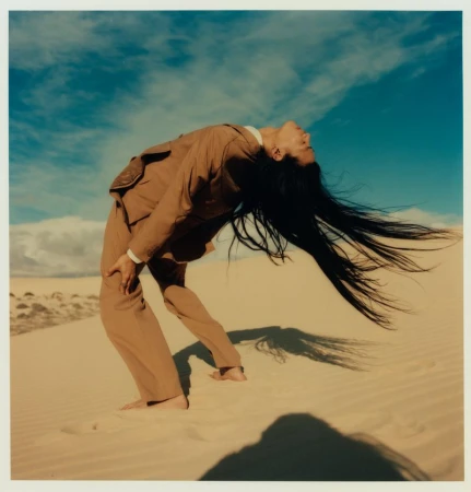 Woman in a tan suit bends backward on sand dunes, head tilted back and long black hair flowing behind her under a blue sky.