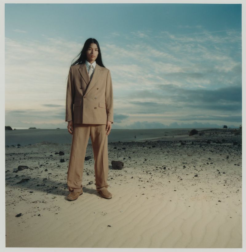 An Asian man stands on a sandy plain in a tan double-breasted suit. He poses in front of a pale blue sky with wispy white clouds.