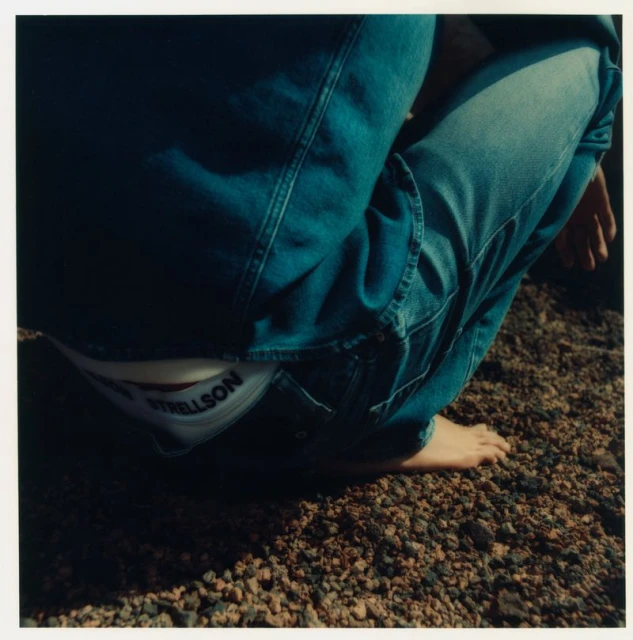 Young adult crouching on rocky ground wearing blue jeans and white underwear; underwear brand visible; bare feet resting on the ground.