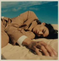 Model lying on sandy terrain wearing a tan suit jacket against a clear blue sky background