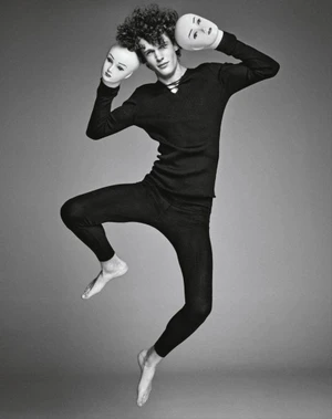 Man with curly hair wearing a black long-sleeve top and leggings jumping while holding two white theatrical masks
