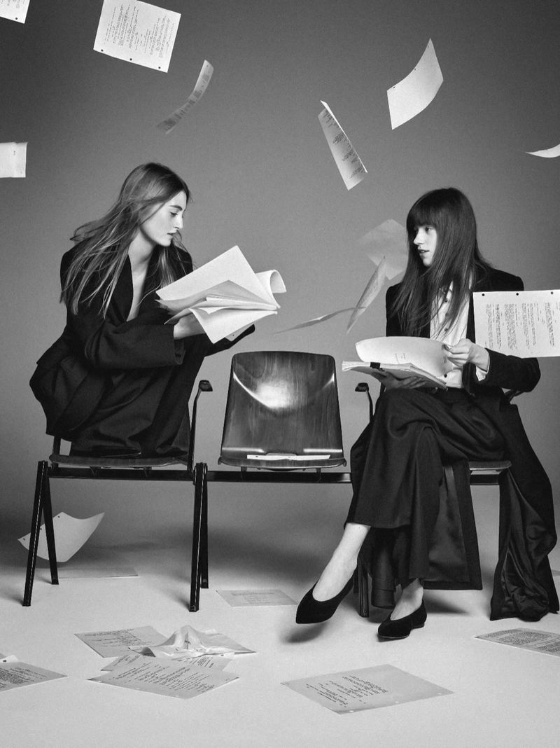Two women sitting on chairs in a studio with papers floating in the air and scattered on the floor around them