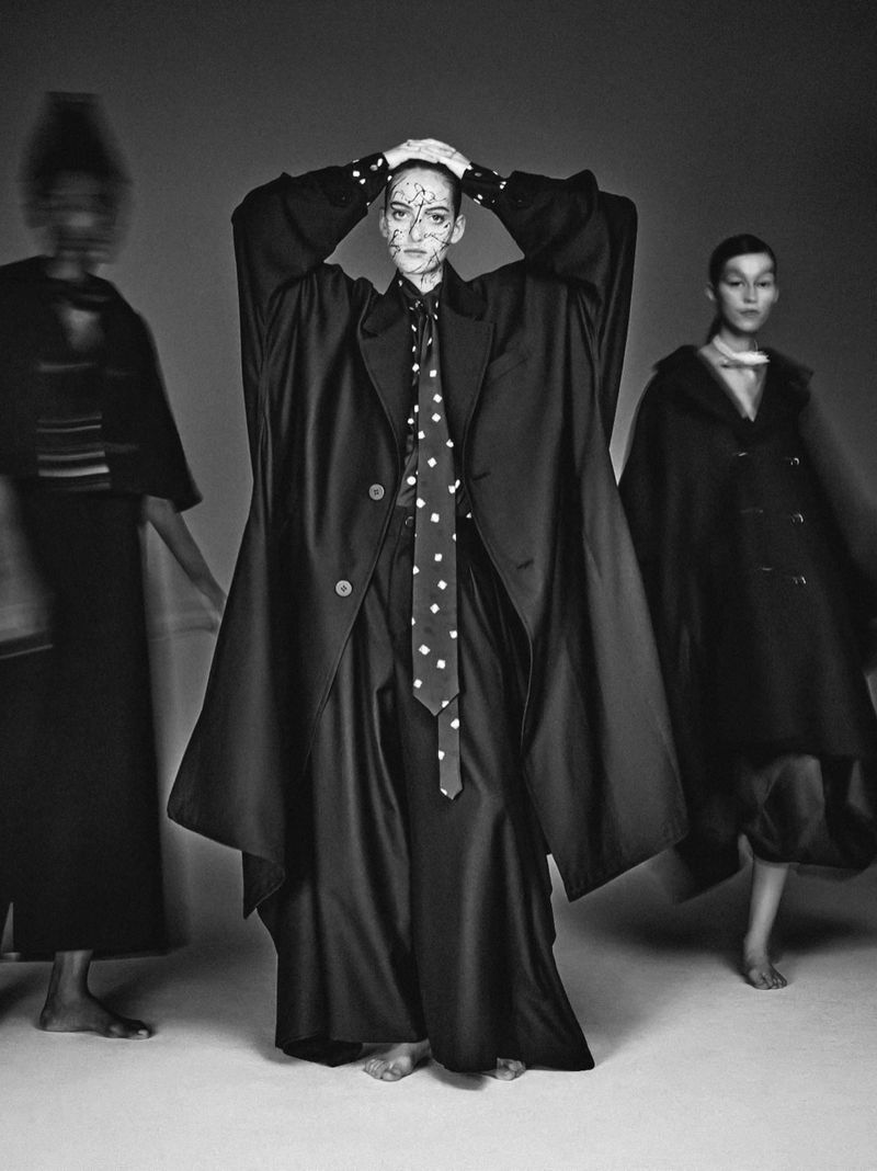Monochrome fashion editorial featuring a central model in a black coat and polka-dot tie with two blurred figures in studio.